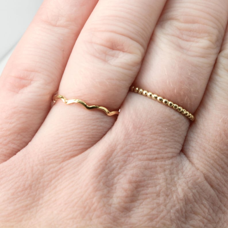 May include: Two gold rings on a finger. One ring is a simple band with small beads, the other ring is a wavy band.