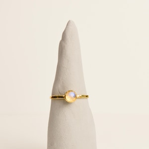 Op de afbeelding: Een gouden ring met een gehamerde band en een kleine, ovale, iriserende edelsteen. De ring wordt getoond op een grijze, kegelvormige standaard. De edelsteen heeft een zachte, parelachtige gloed.