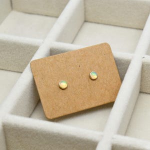 Op de afbeelding: Een paar kleine, ronde oorbellen met opalen op een bruine kaart. De oorbellen zijn gezet in goud.