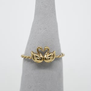 Puede incluir: Un anillo dorado con dos cisnes enfrentados, formando un corazón. El anillo tiene un diseño de banda trenzada. Los cisnes están detallados con plumas texturizadas. El anillo se exhibe sobre un objeto gris en forma de cono.