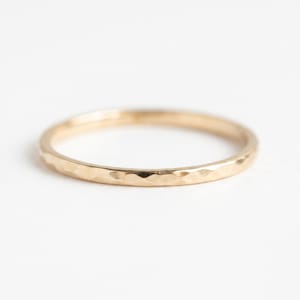 Op de afbeelding: Een dunne, ronde gouden ring met een gehamerd, getextureerd oppervlak dat kleine, reflecterende inkepingen creëert langs de gehele buitenband. Het metaal heeft een warme, gepolijste geeltint en de ring wordt in een eenvoudige stand getoond.