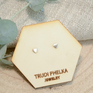 May include: A pair of small, silver heart-shaped stud earrings displayed on a light wooden hexagonal card. The card is branded with the text "TRUDI PHELKA JEWELRY" in a dark brown color. The background includes a neutral-toned fabric and greenery.