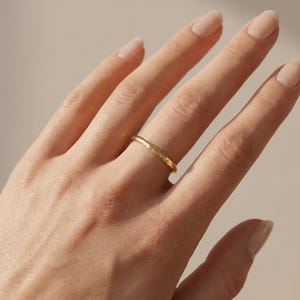 Op de afbeelding: Een dunne gouden ring met een getextureerd, gehamerd oppervlak. De band heeft een onregelmatig, organisch uiterlijk over de gehele omtrek, reflecteert het licht met een metaalachtige glans en behoudt een eenvoudige, cirkelvormige vorm.