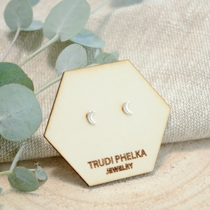 Op de afbeelding: Een paar zilveren halve maan oorbel studs op een houten display kaart met de tekst "Trudi Phelka Jewelry".