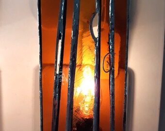 May include: A stained glass nightlight shaped like a small house with a lit bulb inside. The house is made of amber glass with black metal framing.