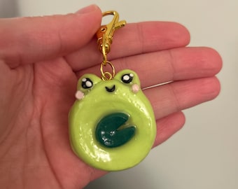 May include: A lime green frog-shaped keychain with a dark green lily pad design. The frog has black eyes, pink cheeks, and a small smile. It is attached to a gold-colored clasp.