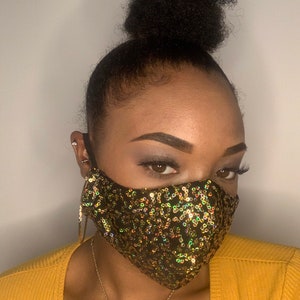 Puede incluir: Una mujer con una máscara facial de lentejuelas negras y doradas. La máscara tiene una forma geométrica y está hecha de pequeñas lentejuelas iridiscentes.