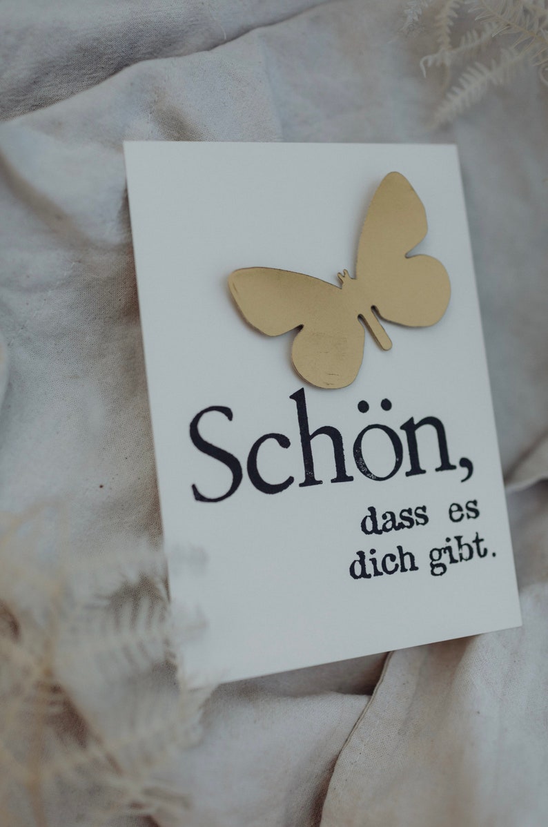 May include: A white card with a gold butterfly cutout and the text "Sch&ouml;n, dass es dich gibt." in black lettering.