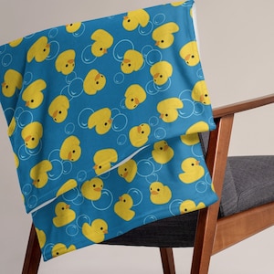 May include: A blue throw blanket featuring a pattern of yellow rubber ducks and white bubbles. The blanket is draped over a wooden chair with a grey cushion.