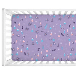 May include: A purple fitted crib sheet with a white crib frame. The sheet has a repeating pattern of pink and blue beakers, atoms, and microscopes.