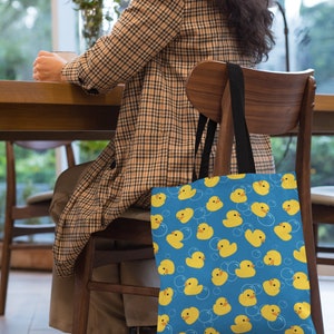 May include: A blue canvas tote bag with a pattern of yellow rubber duckies and white bubbles. The bag has black handles and is hanging on the back of a wooden chair.