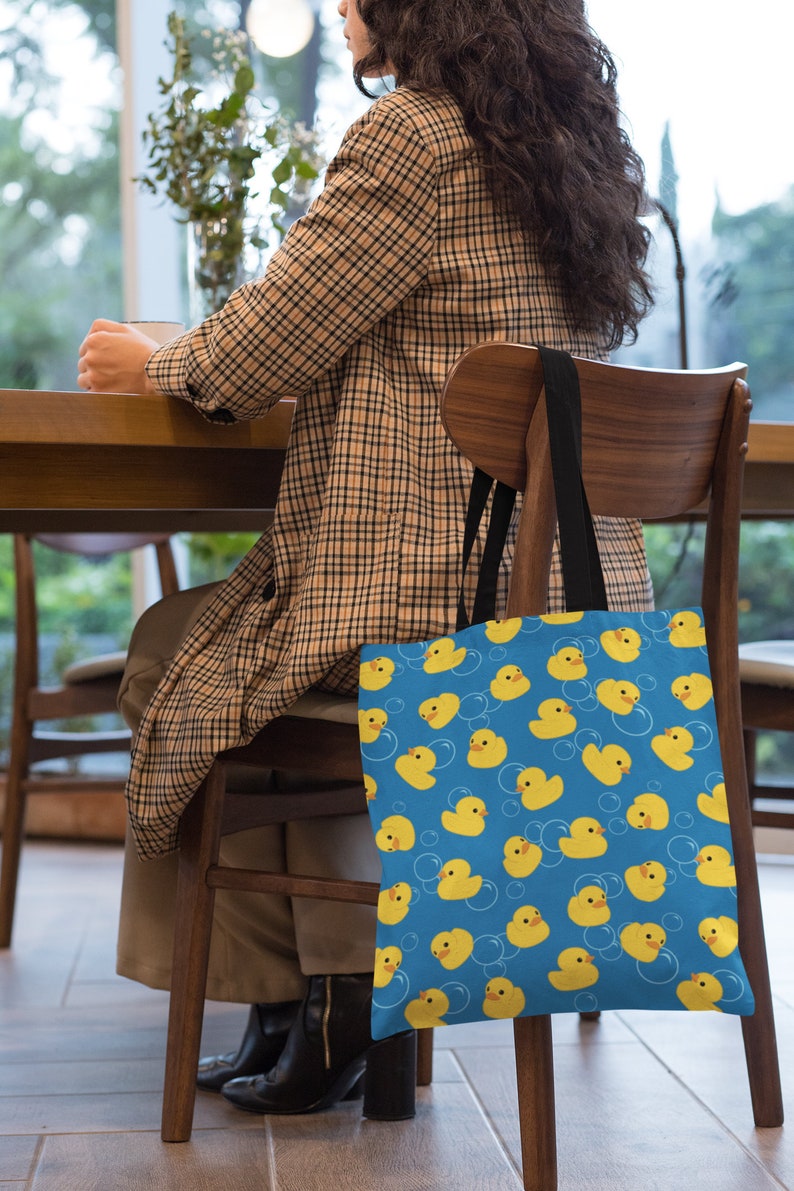 May include: A blue canvas tote bag with a pattern of yellow rubber duckies and white bubbles. The bag has black handles and is hanging on the back of a wooden chair.