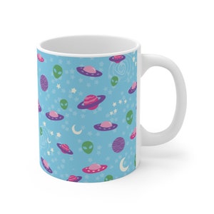 K&ouml;nnte beinhalten: Eine wei&szlig;e Keramiktasse mit blauem Hintergrund und einem sich wiederholenden Muster aus gr&uuml;nen Au&szlig;erirdischen, rosa und violetten Planeten und fliegenden Untertassen. Die Tasse hat einen wei&szlig;en Henkel.