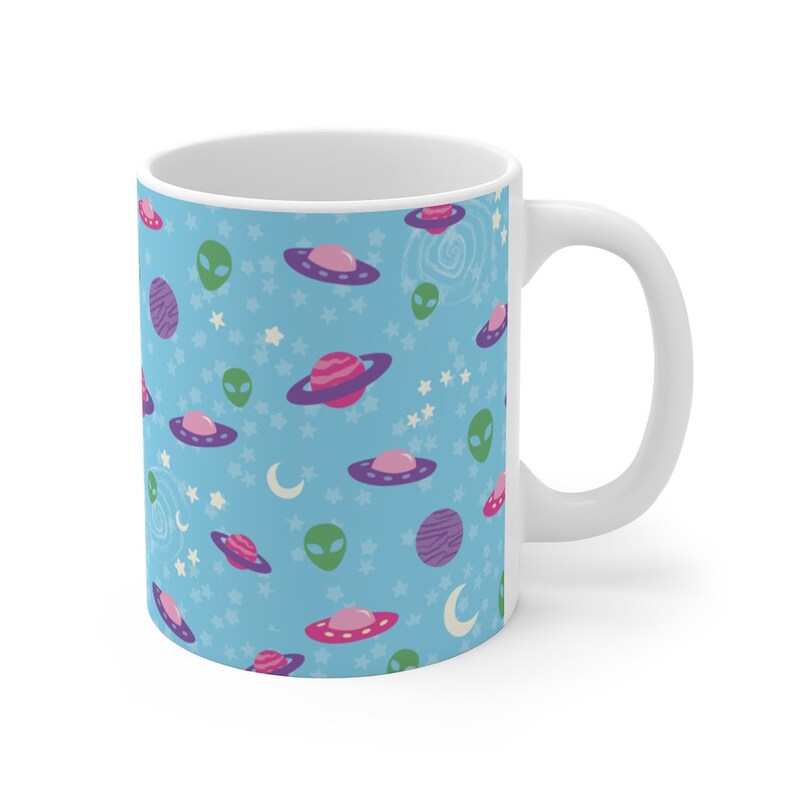 K&ouml;nnte beinhalten: Eine wei&szlig;e Keramiktasse mit blauem Hintergrund und einem sich wiederholenden Muster aus gr&uuml;nen Au&szlig;erirdischen, rosa und violetten Planeten und fliegenden Untertassen. Die Tasse hat einen wei&szlig;en Henkel.