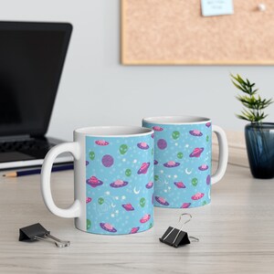 K&ouml;nnte beinhalten: Zwei wei&szlig;e Keramikbecher mit blauem Hintergrund und einem Muster aus rosa und violetten Planeten, gr&uuml;nen Au&szlig;erirdischen und wei&szlig;en Sternen. Die Becher stehen auf einer hellbraunen Holzoberfl&auml;che.