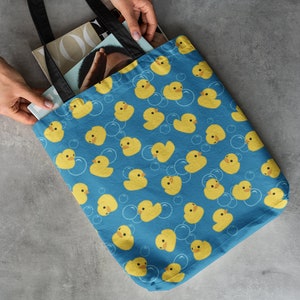 May include: A blue tote bag with a pattern of yellow rubber duckies and white bubbles. The bag has black handles and is filled with magazines.