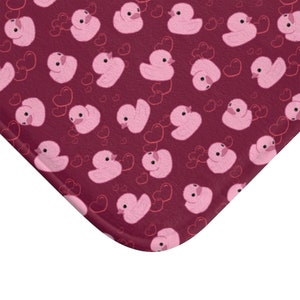 May include: A burgundy bath mat with a repeating pattern of pink rubber ducks and hearts.