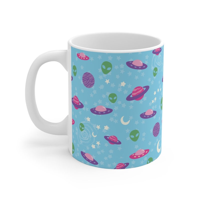 K&ouml;nnte beinhalten: Eine wei&szlig;e Keramiktasse mit blauem Hintergrund und einem sich wiederholenden Muster aus gr&uuml;nen Au&szlig;erirdischen, rosa und violetten Raumschiffen und Planeten.