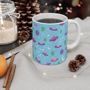 K&ouml;nnte beinhalten: Eine blaue Keramiktasse mit einem Weltraum-Motiv. Die Tasse zeigt Aliens, Planeten, Sterne und Monde in Rosa, Lila und Gr&uuml;n. Die Tasse ist mit Kaffee gef&uuml;llt.