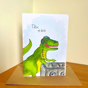 Puede incluir: Tarjeta de felicitación con un T-Rex verde con auriculares y operando mesas de DJ. La tarjeta tiene el texto "T-Rex on decks" en escritura negra. La tarjeta está dentro de un sobre marrón, sobre una superficie de madera.