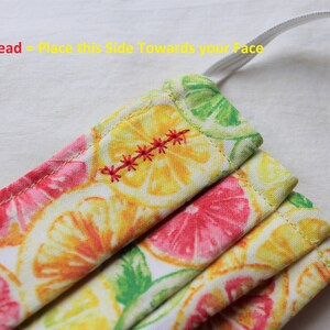 May include: A white face mask with a lemon and grapefruit print. The mask has a red thread sewn across the top, indicating the side to wear facing the wearer's face. The text "Red Thread - Place this Side Towards your Face" is visible at the top of the image.