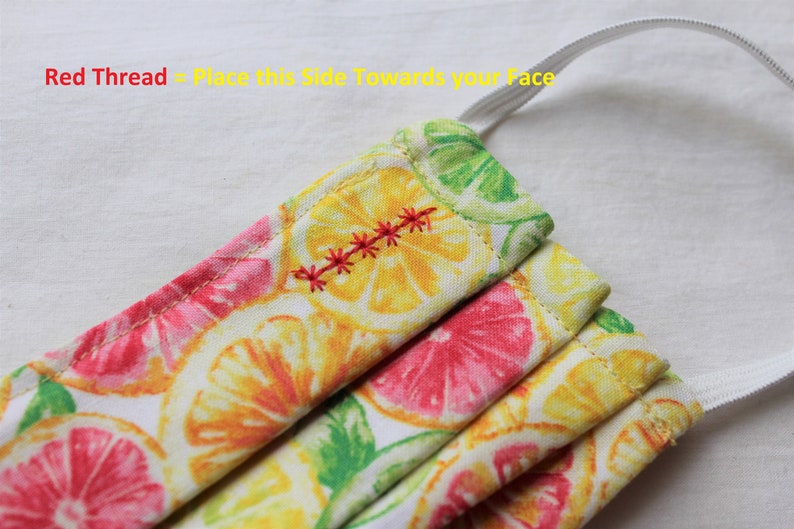 May include: A white face mask with a lemon and grapefruit print. The mask has a red thread sewn across the top, indicating the side to wear facing the wearer's face. The text "Red Thread - Place this Side Towards your Face" is visible at the top of the image.