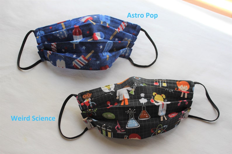May include: Two fabric face masks with black elastic ear loops. The first mask has a blue background with red, white, and blue popsicle designs and the text "Astro Pop". The second mask has a black background with a science theme and the text "Weird Science".