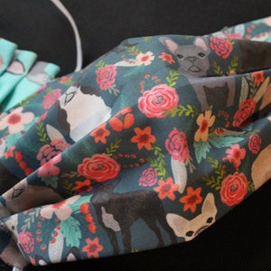 May include: Close-up of several fabric face masks. The masks feature a teal and dark teal floral pattern with images of French bulldogs. The masks have white elastic ear loops.
