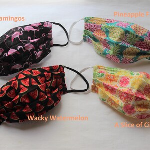 May include: Four different fabric face masks with various patterns. The masks are pleated and have black elastic ear loops. The patterns are: "Dancing Flamingos" with pink flamingos on a black background, "Pineapple Frenzy" with yellow pineapples on a teal background, "Wacky Watermelon" with red watermelon slices on a black background, and "A Slice of Citrus" with yellow citrus slices on a white background.