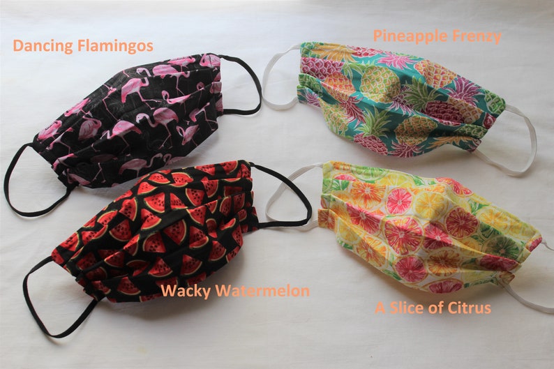 May include: Four different fabric face masks with various patterns. The masks are pleated and have black elastic ear loops. The patterns are: "Dancing Flamingos" with pink flamingos on a black background, "Pineapple Frenzy" with yellow pineapples on a teal background, "Wacky Watermelon" with red watermelon slices on a black background, and "A Slice of Citrus" with yellow citrus slices on a white background.