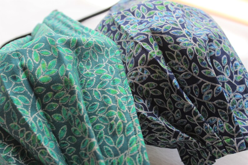 May include: Two face masks with a leafy pattern. One mask is teal with green leaves and the other is navy blue with green leaves.