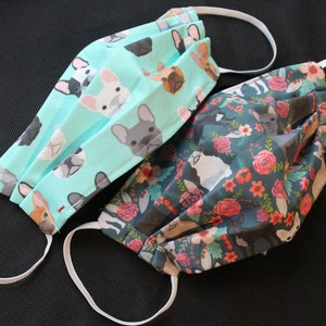 May include: Two face masks with white elastic ear loops. One mask is light teal with a pattern of French bulldog faces. The other mask is dark teal with a floral and French bulldog pattern. Both masks are made of fabric.