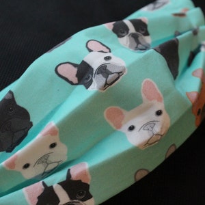May include: A pleated face mask featuring a repeating pattern of French bulldog faces in various colors on a light teal background. The dogs have black, white, and brown fur. The mask has white elastic ear loops.