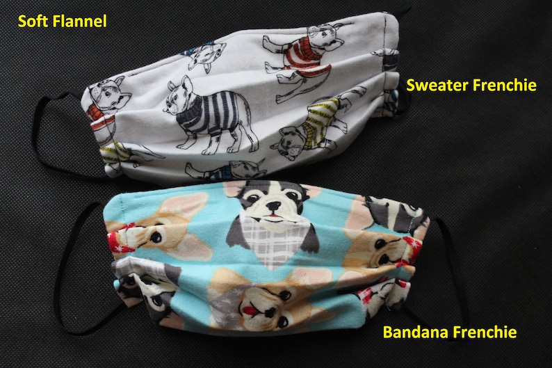 May include: Two face masks with black elastic ear loops. One mask is white with French bulldog illustrations wearing sweaters. The other is light blue with French bulldog illustrations wearing bandanas. Text on the masks reads "Soft Flannel", "Sweater Frenchie", and "Bandana Frenchie".