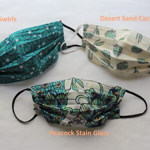 May include: Three different fabric face masks with black elastic ear loops. The masks are folded and feature different patterns: teal with white stars and swirls, beige with green cactus plants, and white with a peacock feather design. The masks are labeled "Stars N Swirls", "Desert Sand Cactus", and "Peacock Stain Glass".