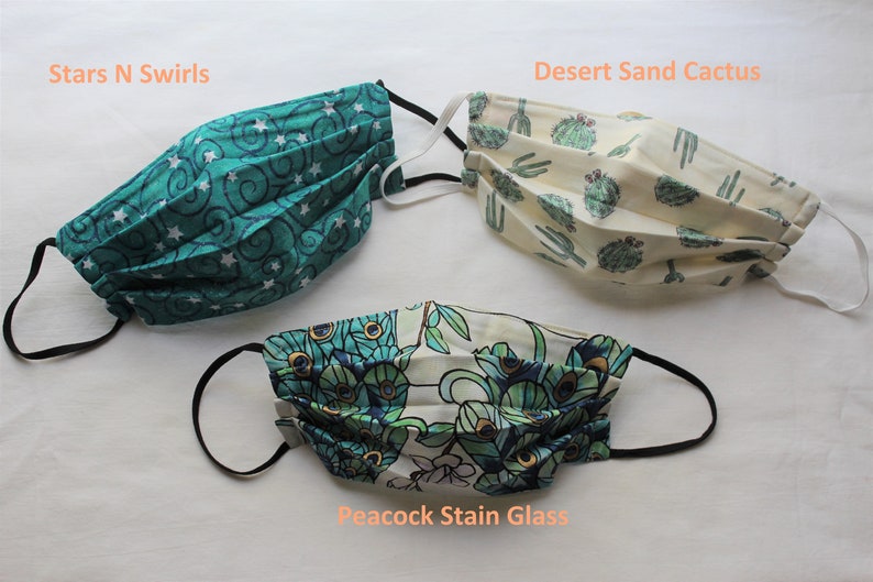 May include: Three different fabric face masks with black elastic ear loops. The masks are folded and feature different patterns: teal with white stars and swirls, beige with green cactus plants, and white with a peacock feather design. The masks are labeled "Stars N Swirls", "Desert Sand Cactus", and "Peacock Stain Glass".