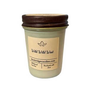 May include: A glass jar candle with a brown lid and a beige label. The label reads "Wild Wild West" with a floral design and the website Rockridgecandles.com. The candle is filled with a light green wax.