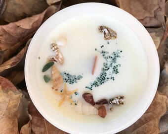 May include: A white candle with a variety of seashells, starfish, and small stones embedded in the wax. The candle is in a clear glass jar.