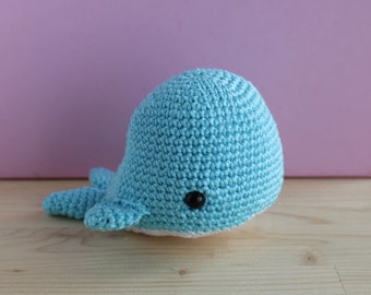 May include: A light blue crocheted whale toy with a white belly and black eyes. The whale is sitting on a light brown wooden surface.