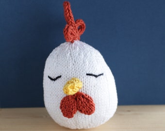 May include: A white knitted chicken toy with a red comb and wattle, and yellow beak. The chicken has closed eyes, giving it a sleepy appearance. The background is a dark blue colour.