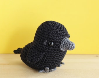 May include: A black crocheted crow with gray beak and black eyes. The crow is sitting on a wooden surface.