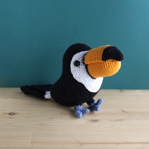 Puede incluir: Un tucán de ganchillo hecho a mano con un gran pico naranja y negro, cuerpo negro, detalles blancos y patas azules. El ave está posada sobre una superficie de madera contra un fondo turquesa. Esta escultura blanda es un artículo decorativo único.