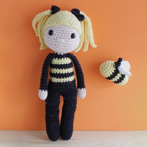 May include: A handmade crochet doll with a bee theme. The doll has a light yellow head with blonde pigtails and black bows. The body is black with yellow stripes, and the doll is wearing black pants. A separate crochet bee is also present.