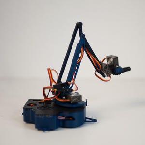 Op de afbeelding: Een kleine robotarm in donkerblauw en zwart, met oranje bedrading. Gemonteerd op een donkerblauwe basis. Het geheel staat op een witte ondergrond, wat doet denken aan een model of educatief hulpmiddel.