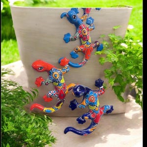 May include: Three ceramic lizard figurines attached to the side of a large, light-colored planter. Each lizard features a glossy finish with vibrant, multi-colored floral and geometric patterns in shades of red, blue, yellow, and green.