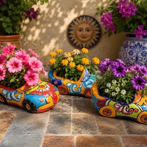 May include: Three ceramic car-shaped planters featuring vibrant, hand-painted patterns in blue, yellow, red, and green. Each planter holds a different variety of flowers: pink petunias, yellow lantanas, and purple petunias with small white blossoms.