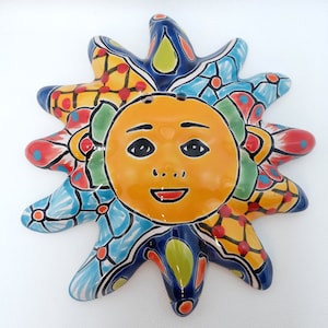 May include: A ceramic sun wall plaque featuring a central yellow face with black eyes and a smiling mouth. The sun rays are decorated with vibrant patterns in blue, yellow, green, and red, including floral motifs, geometric grids, and solid color accents.