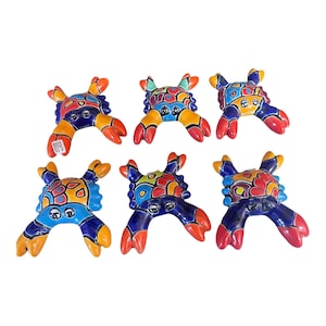 May include: Six ceramic crab figurines arranged in two rows. Each crab features a glossy finish with hand-painted, vibrant patterns in shades of blue, orange, red, yellow, and green. The crabs have distinct eyes and large claws, with one crab displaying a small white label.