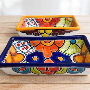 May include: Two rectangular ceramic dishes featuring hand-painted floral patterns in vibrant shades of orange, blue, red, yellow, and green. The front dish has a deep blue rim, while the back dish features a bright orange rim.