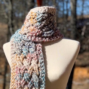 May include: A long, hand-crocheted scarf featuring a complex cable-knit pattern. The yarn is a variegated blend of cream, tan, soft blue, and muted red tones. The scarf ends in a straight fringe of matching yarn strands hanging from the bottom edge.
